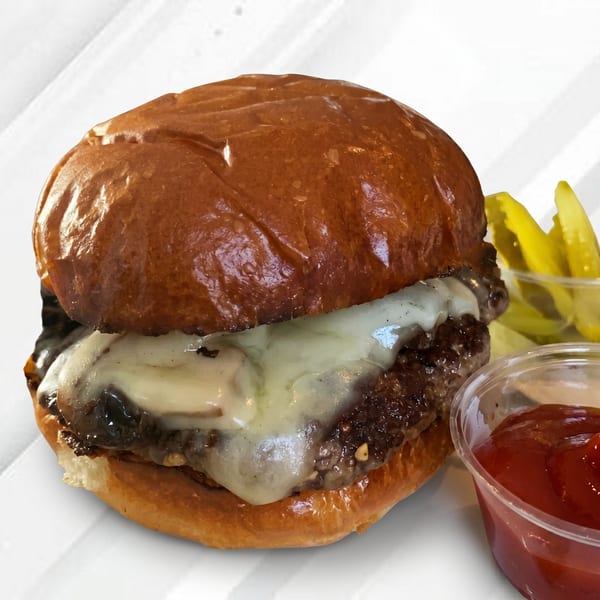 Cheeseburger with melted white cheese, a side of pickles, and ketchup in a clear cup.