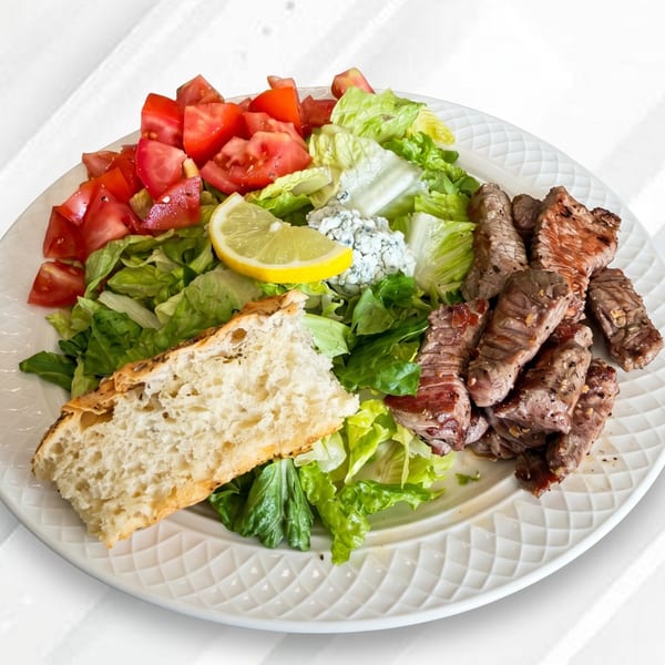 Grilled steak pieces, fresh green salad with tomatoes, blue cheese, lemon, and a slice of bread.