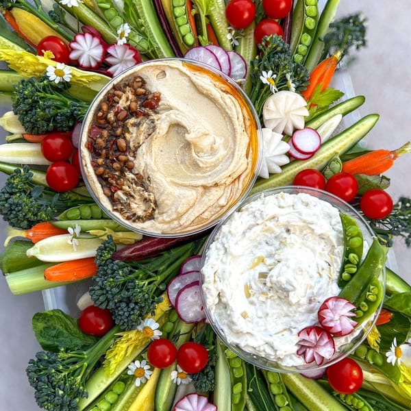 Colorful vegetable platter with two dips, including hummus, surrounded by fresh radishes, tomatoes, and broccoli.