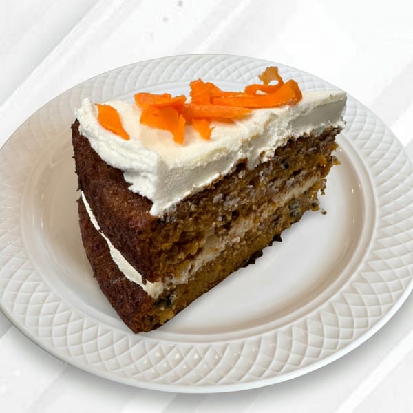 A slice of carrot cake with white cream cheese frosting and candied carrot curls on a white plate.
